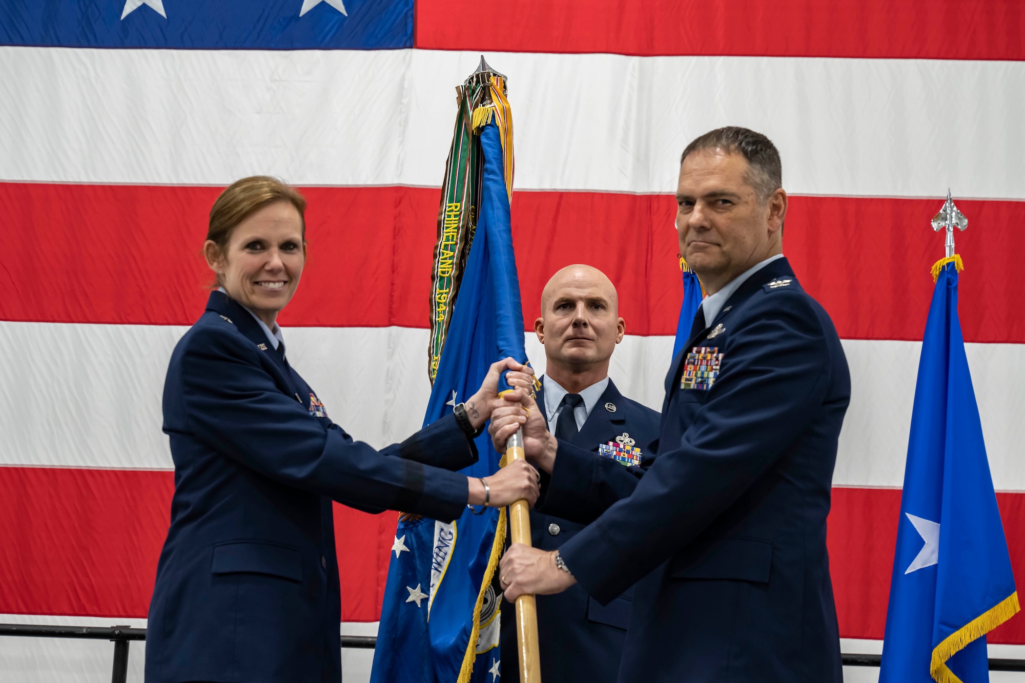 A woman hands a man a flag representing command of the 442d Fighter Wing while another man stands at attention in the background. All three are wearing U.S. Air Force service dress and standing in front of an enormous American flag.