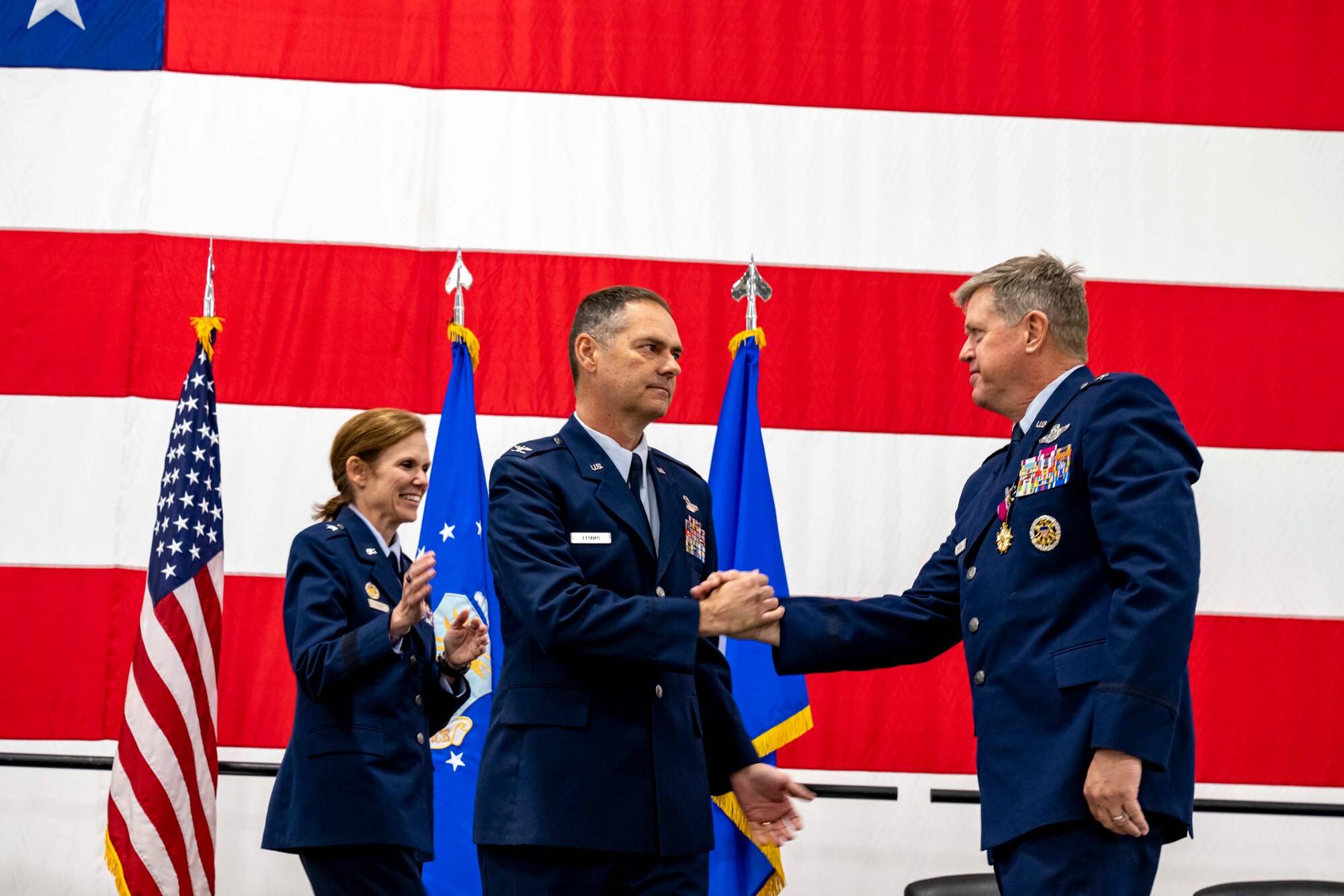 Two men in U.S. Air Force service dress shake hands in the foreground while a similarly-attired woman smiles behind them all in front of a very large American flag.