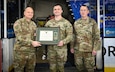 Three soldiers in uniform pose for a photo with a framed certificate and medal.
