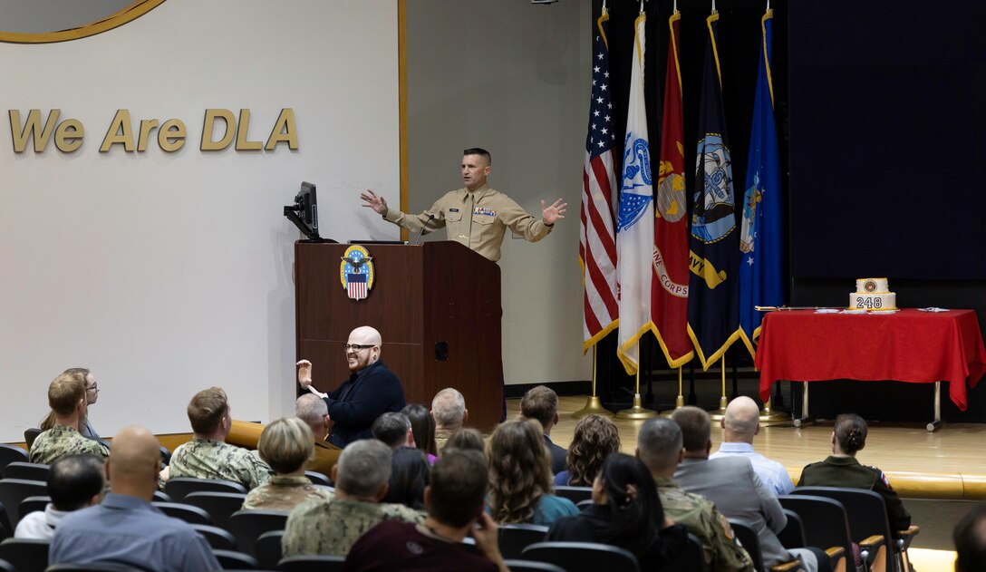 A Marine stretches his arms out while speaking at a podium on a stage in a large auditorium.