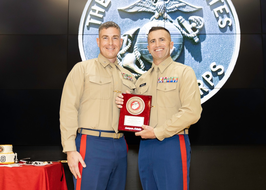 Two smiling Marines hold a certificate on a auditorium stage. They are wearing khaki uniforms with blue pants and have buzz cuts.