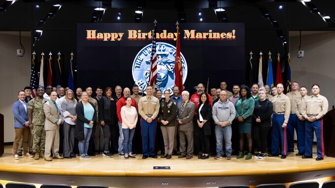 Large group of people stand on stage with a Happy Birthday Marines in the background.