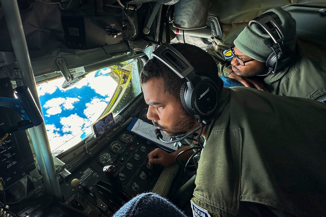 Two airmen wearing headsets look at the sky through a window on an airborne aircraft.