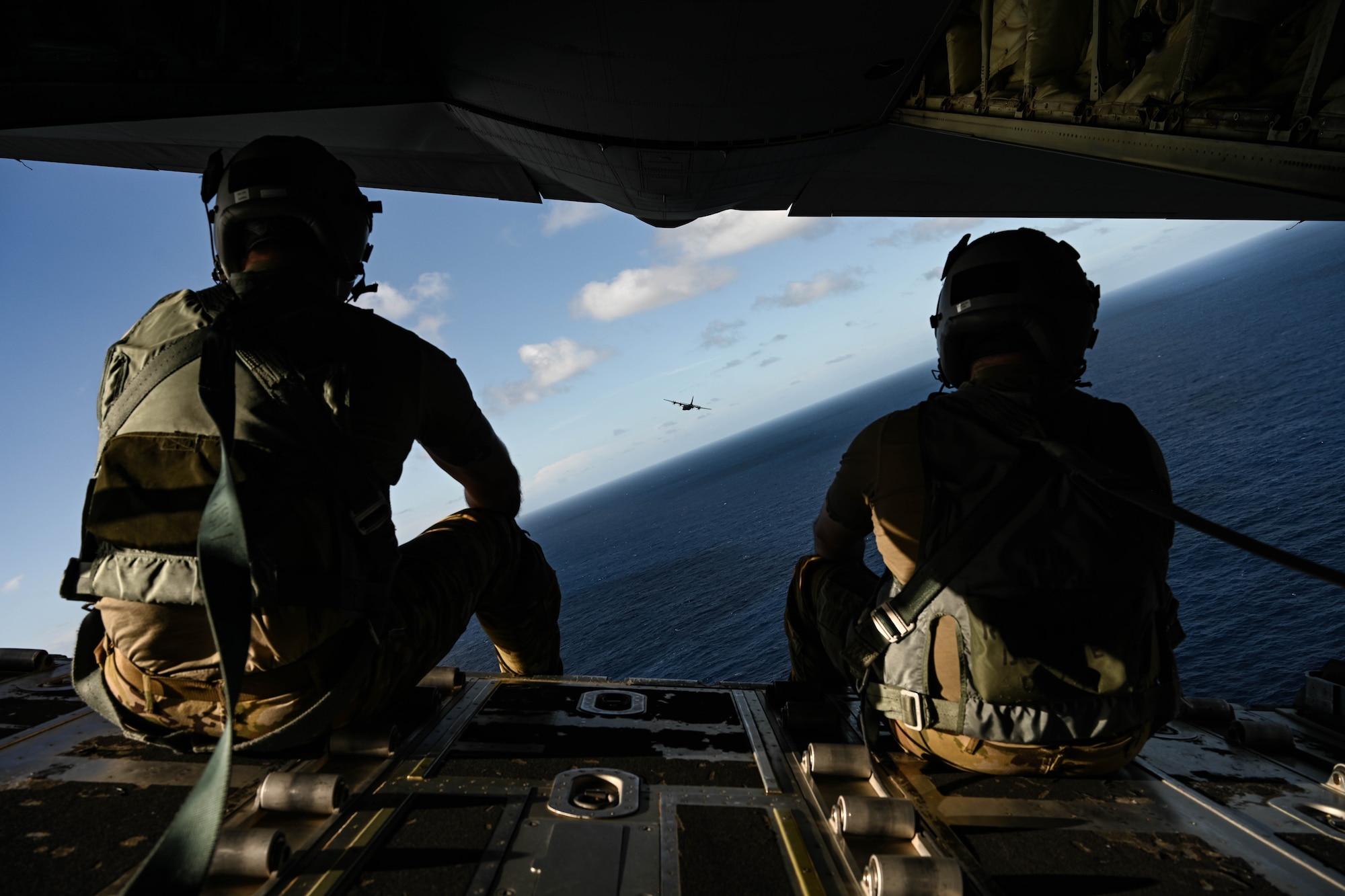 Two men in uniform watch a military aircraft fly over the ocean