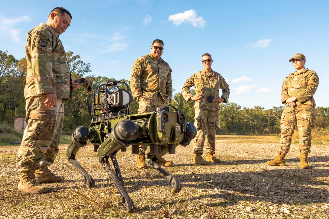 Airmen gather around a robot dog as it walks on a rocky surface in a field.