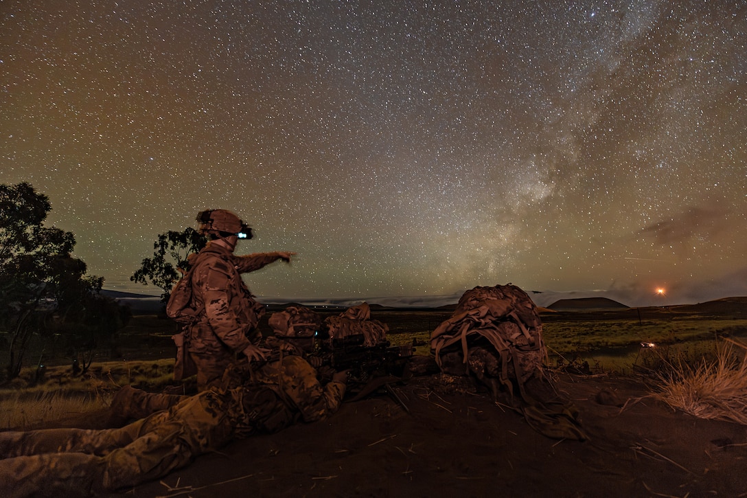 A soldier kneels next to a fellow soldier laying on the ground while pointing to an explosion in the distance under a starry sky.