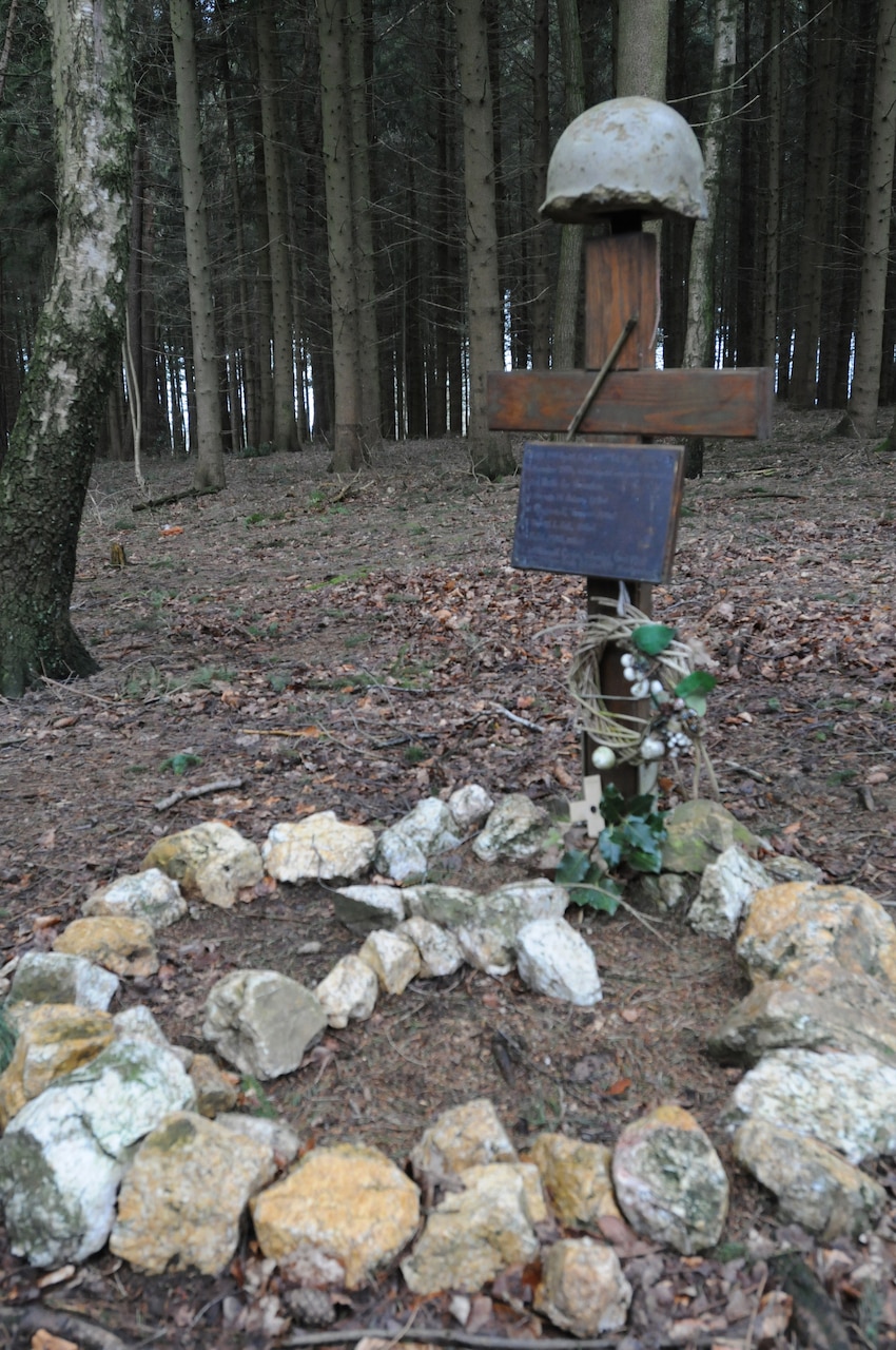 A circle of rocks surrounds a gravesite cross with a plaque and a helmet on top of it in a section of woods.