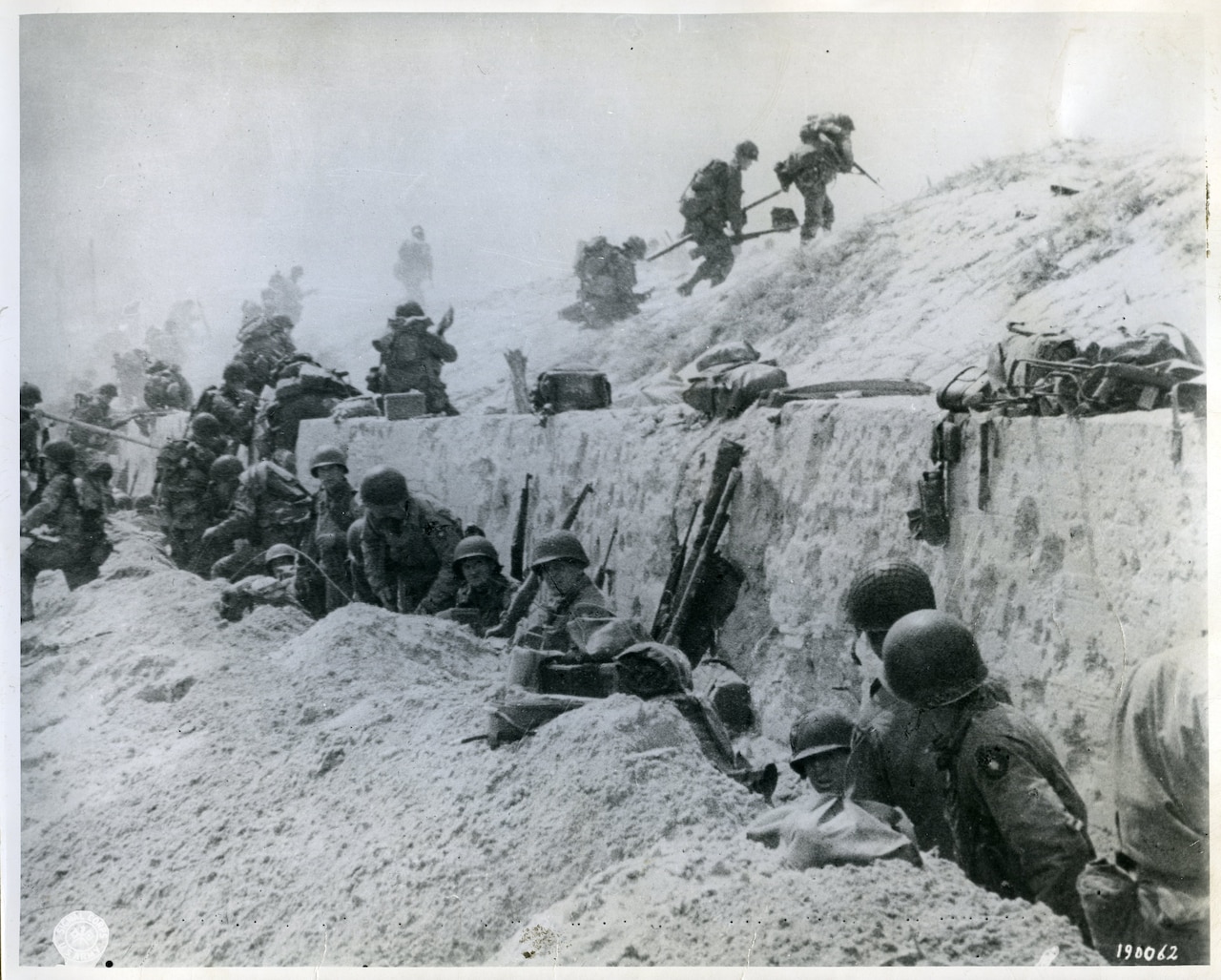 Several men duck down around a sand ledge. Others are climbing it.