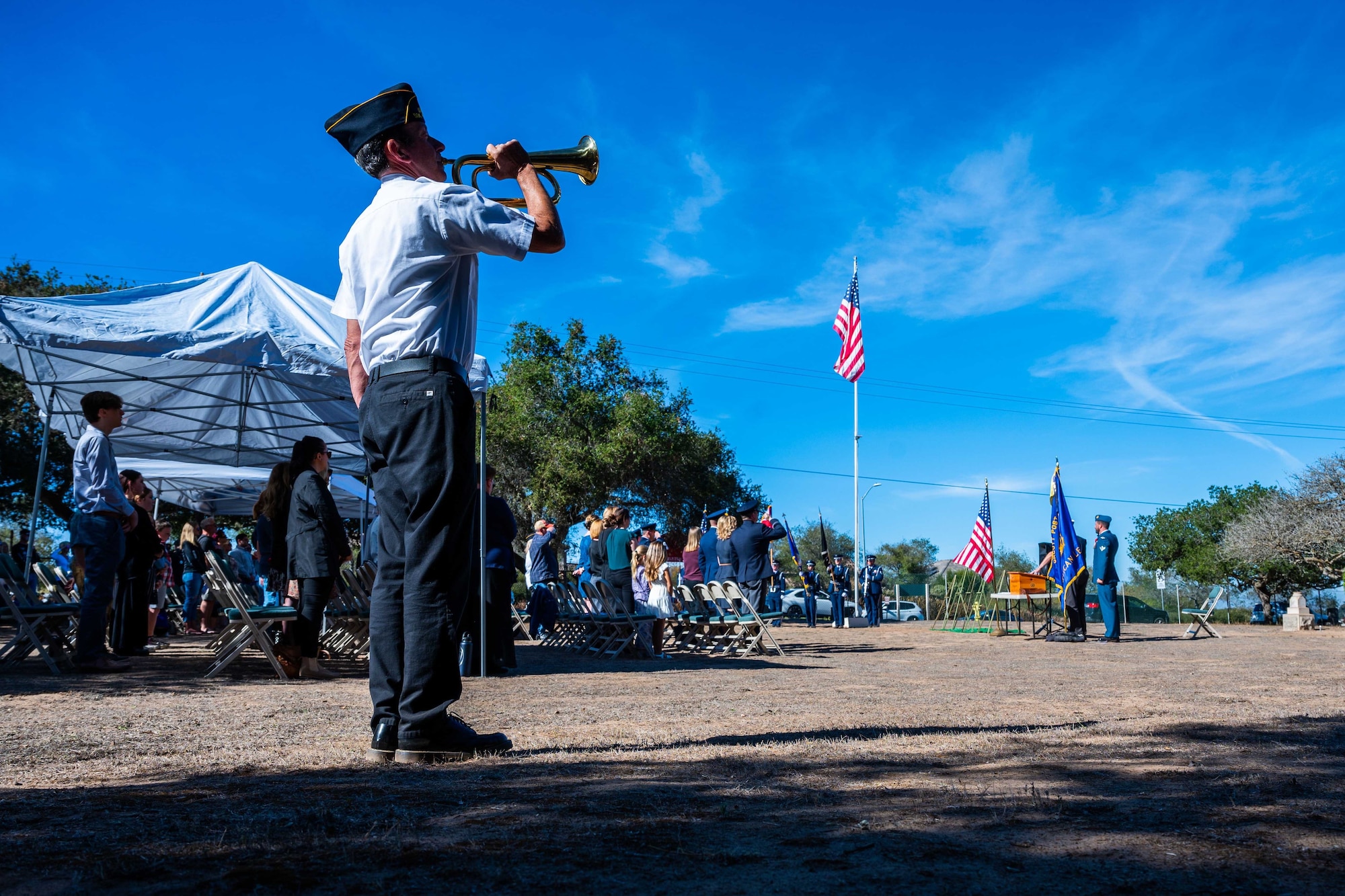 A man plays the bugle in an open field to attendees of a ceremony.