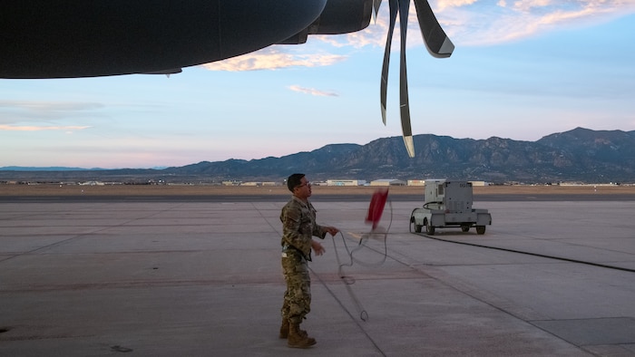 A man in uniform with C-130 propellers overhead pulls down a red object attached to a string.