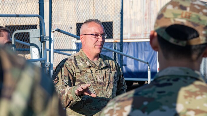 A man in uniform holds his arm out while speaking to a group of other people in uniform.