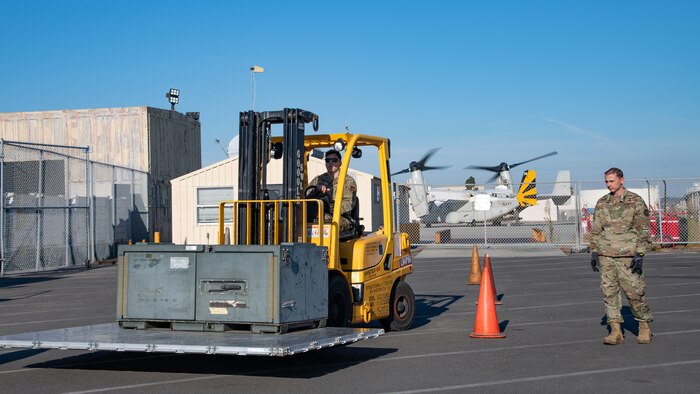 A man in uniform stands nearby as another man in uniform drives a forklift carrying a pallet with a crate on it.