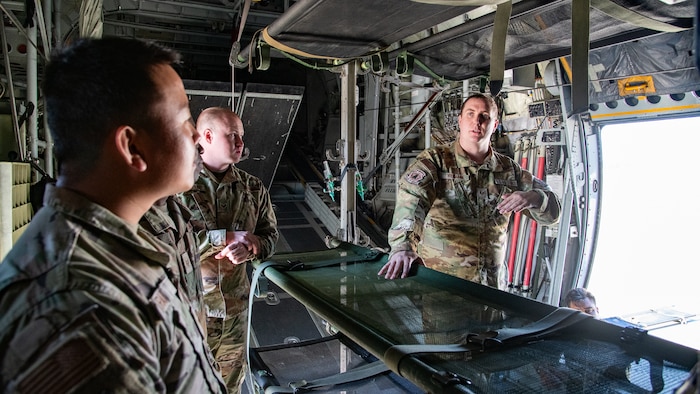 A man in uniform stands on one side of a litter talking to another man in uniform on the other side of it aboard a C-130 aircraft cargo bay.