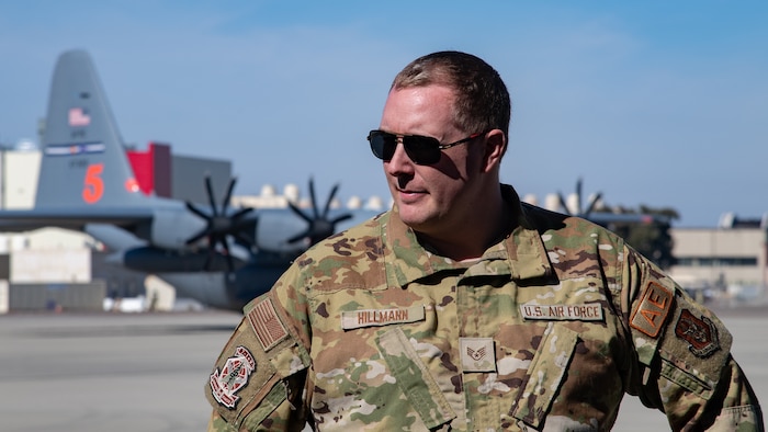A man in uniform wearing sunglasses looks off to the side with a parked C-130 aircraft in the background.