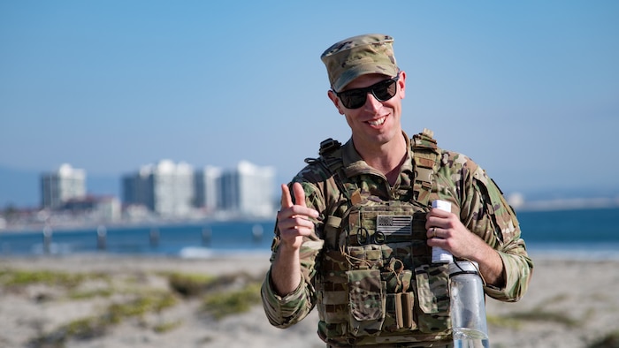 A man in uniform with a San Diego pier in the background points at someone off-camera while smiling.