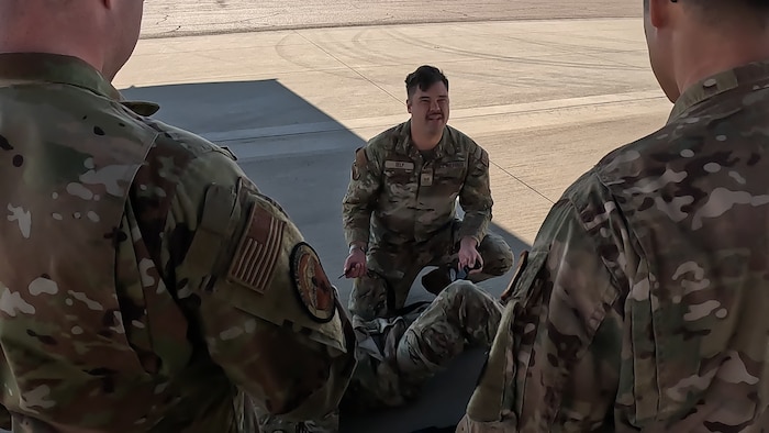 A man in uniform kneels in front of another man in uniform laying down with a tourniquet around his leg. The first man is looking up speaking to onlookers.