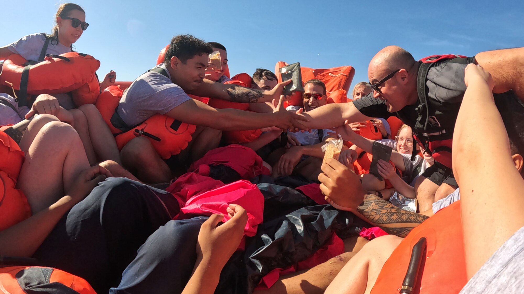 A man reaches out to grasp another man's arm in a life raft in the ocean surrounded by other people.