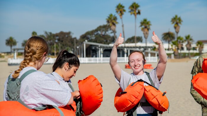 A woman wearing a life raft raises two arms in thumbs up on a beach with two other women nearby also wearing life rafts.