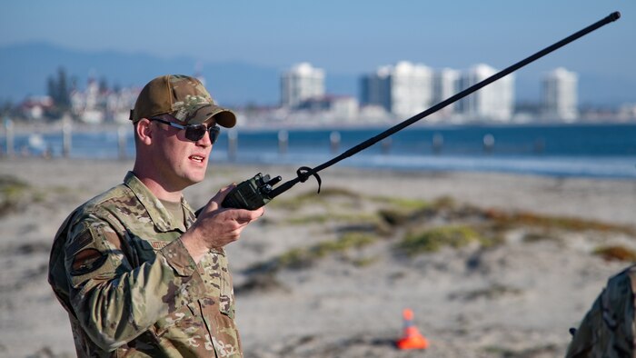 A man in uniform on a beach holds a radio with a long antenna. A pier in San Diego is in the background.