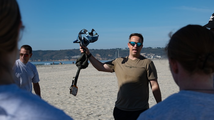 A man wearing sunglasses on a beach holds up an oxygen mask as multiple people observe.