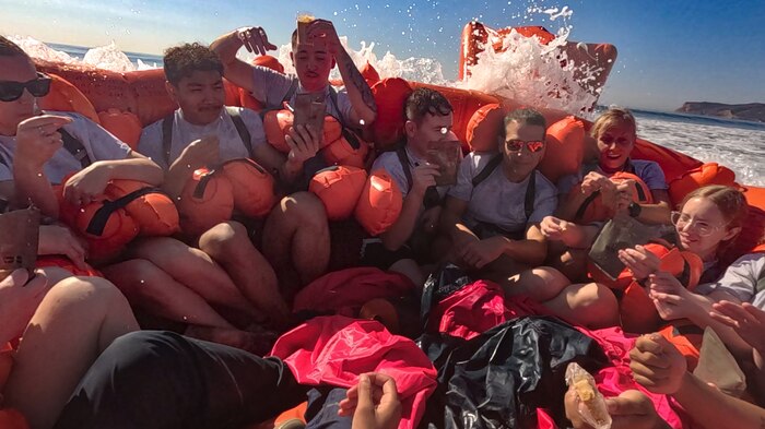 A group of people shoulder-to-shoulder in a life raft are hit by a wave in the ocean.
