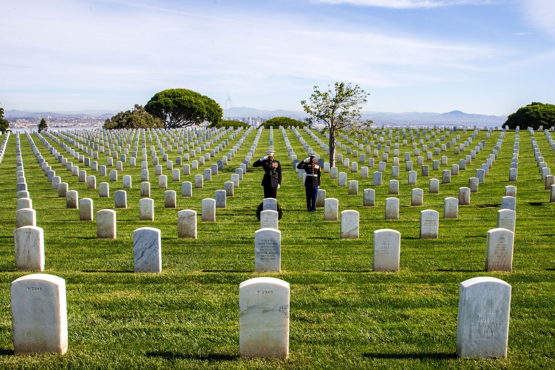 Two Marine Corps officers stand in a large graveyard and salute a headstone.