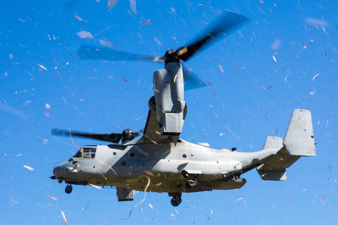 A military aircraft in flight kicks up debris. A bright blue sky can be seen in the background.