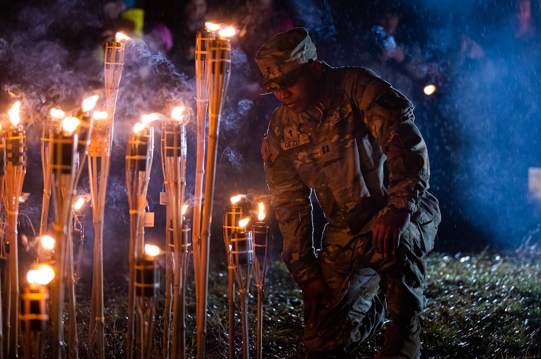 A uniformed service member takes a knee in front of a row of torches at night.