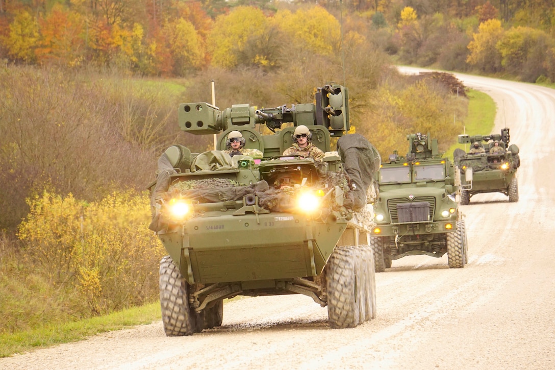 Soldiers ride in armored vehicles on a dirt road surrounded by trees.