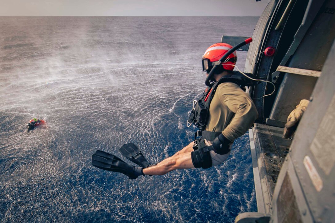 A sailor wearing diving gear jumps from a helicopter into a body of water as a fellow service member treads water to the left.