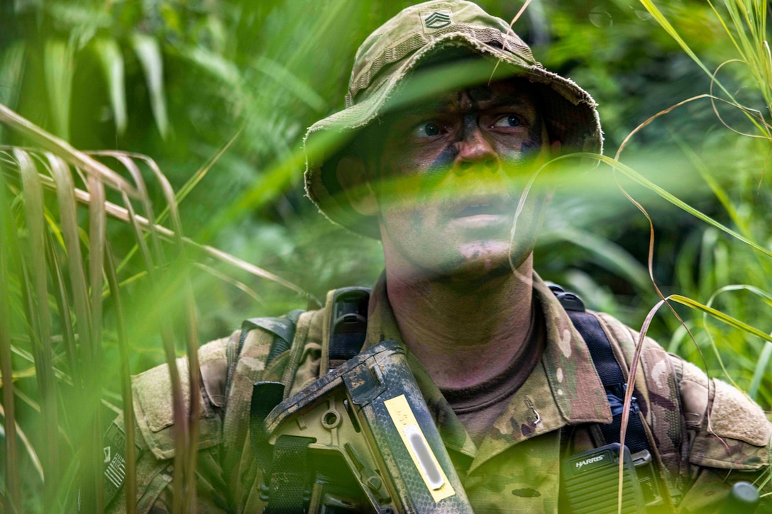 A leaf covers part of a soldier’s camouflage-covered face as they look to the right while standing in the jungle.