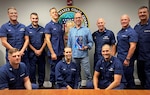 9 uniformed members and 1 civilian stand in front of a United States Coast Guard logo and pose for their photo presenting the award.