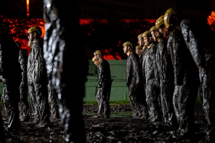 group of men in uniform stand, covered in mud with yellow helmets on their head