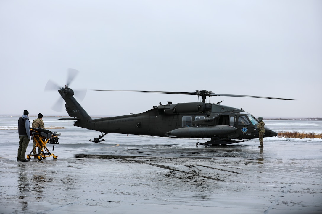 From the left, Alaska Army National Guard Public Affairs Officer Dana Rosso, Joint Force Headquarters, and Chief Warrant Officer 3 Bryan Kruse, Bethel Army Aviation Operating Facility commander, prepare to move an ambulance gurney towards an AKARNG UH-60L Black Hawk helicopter to assist Chief Warrant Officer 3 Nick Lime, an aviation maintenance technician and crew chief with Delta Co., 2-211th (GSAB), safely transporting a critically ill patient during a medical evacuation from Napaskiak to Bethel, Nov. 15, 2023.