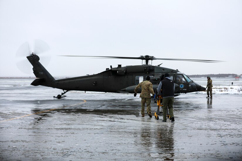 From the left, Alaska Army National Guard Chief Warrant Officer 3 Bryan Kruse, Bethel Army Aviation Operating Facility commander, and Public Affairs Officer Dana Rosso, Joint Force Headquarters, push an ambulance gurney towards an AKARNG UH-60L Black Hawk helicopter to assist Chief Warrant Officer 3 Nick Lime, an aviation maintenance technician and crew chief with Delta Co., 2-211th (GSAB), safely transporting a critically ill patient during a medical evacuation from Napaskiak to Bethel, Nov. 15, 2023.