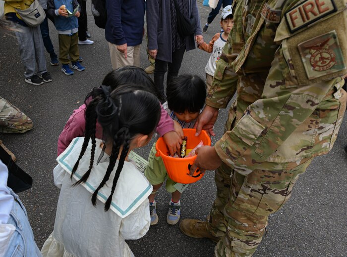 children gather around an orange bucket with candy being handed out by a man in uniform.