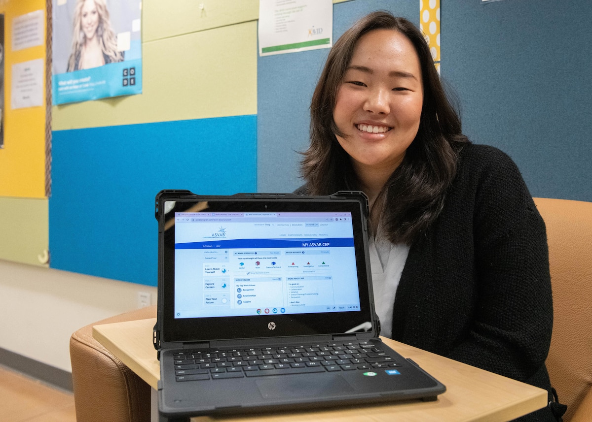 A Highschooler smiles and poses for a photo behind a laptop. the laptop is facing the viewer's direction.