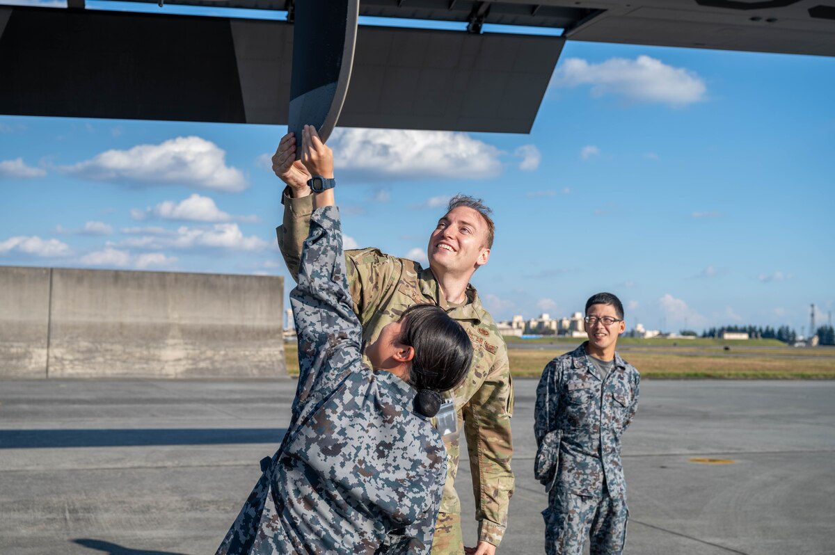 Three people in uniform are in the photo. Two of them are interacting with the propeller of an aircraft while the other person stand adjacent to them watching and smiling.