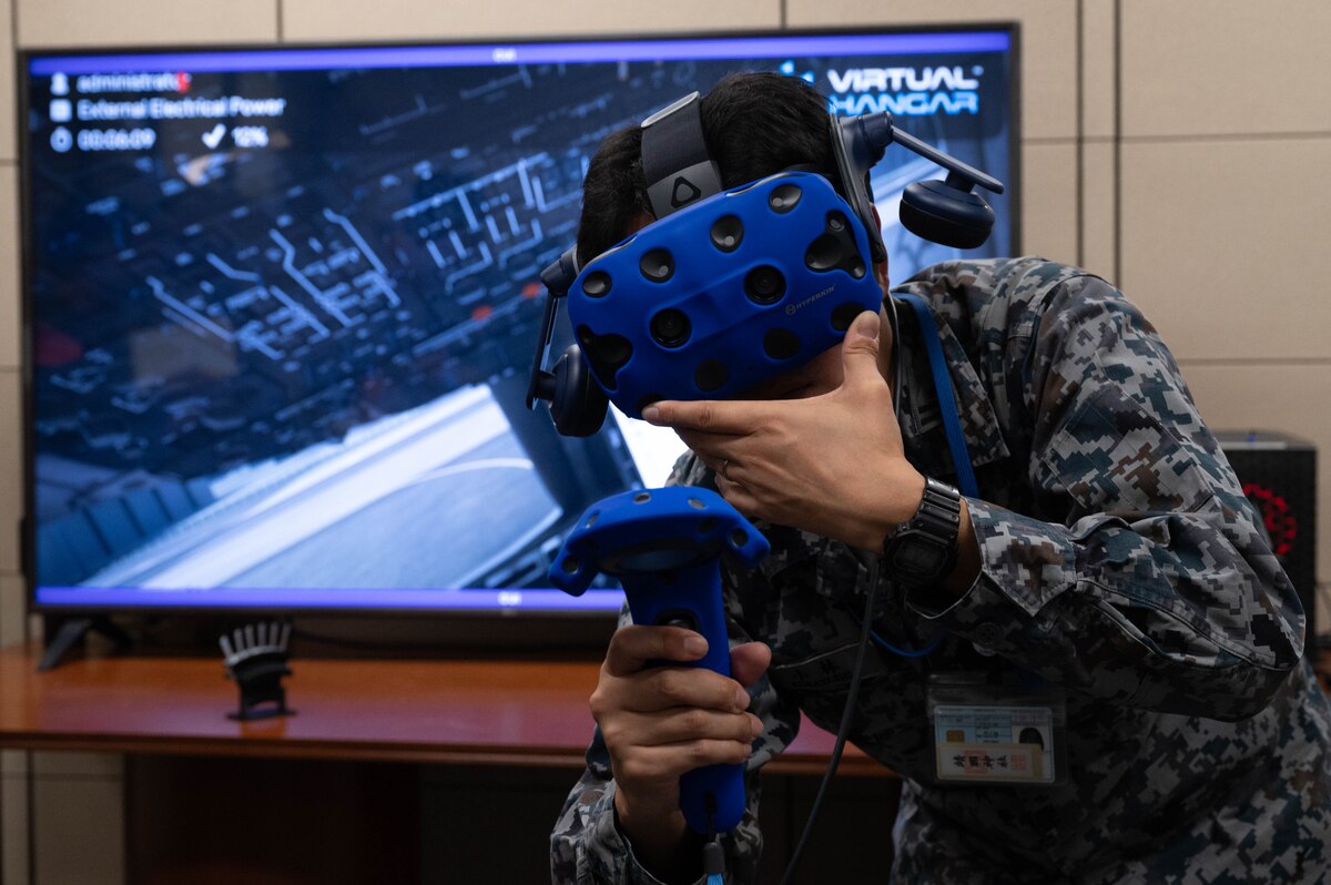 A man in uniform poses wearing a VR headset and holding on to the VR remote with a tv behind him displaying the Virtual cockpit that he sees with the head set on.
