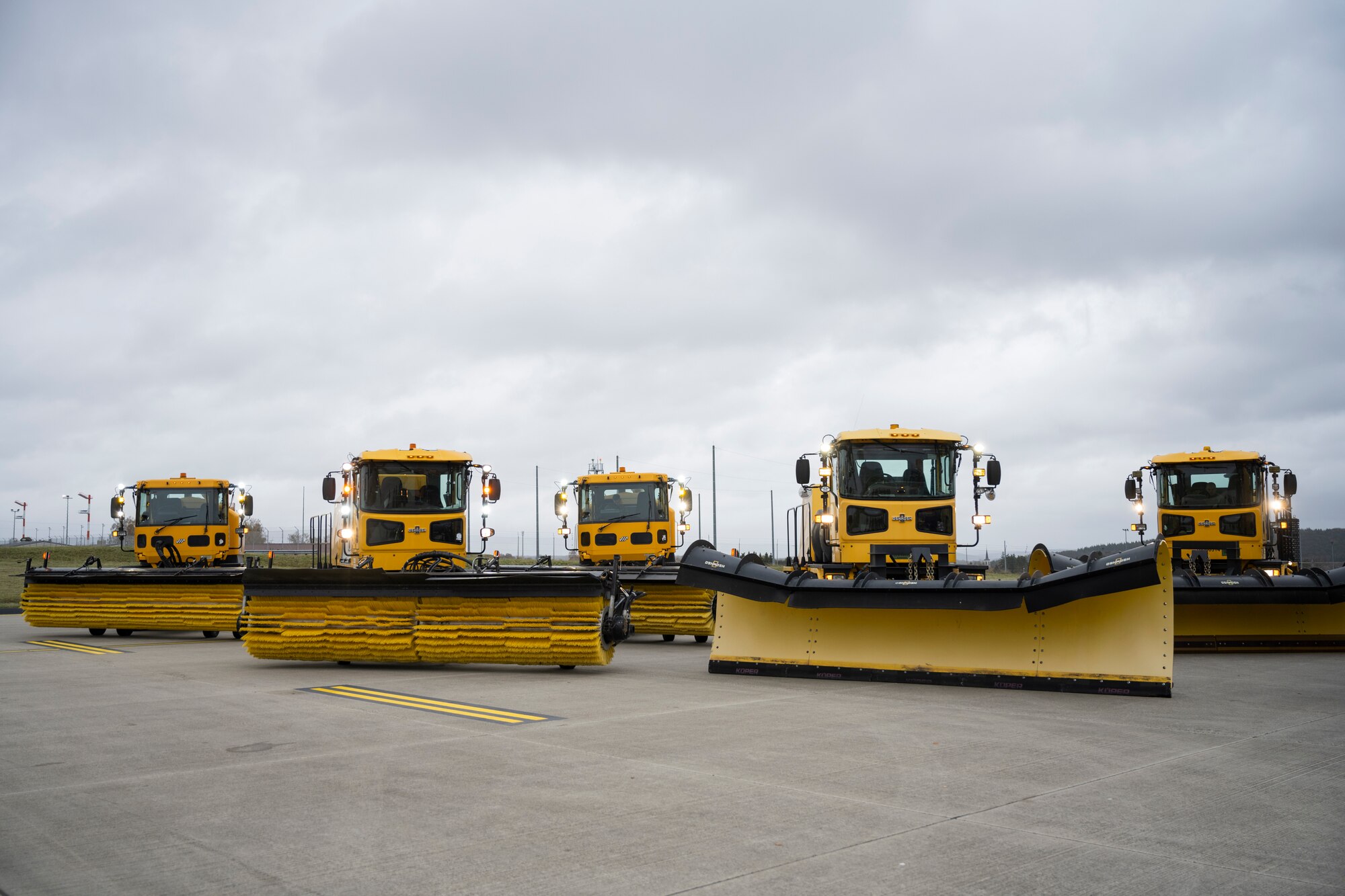 Five large snow removal vehicles are parked on a concrete pad.