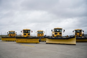 Five large snow removal vehicles are parked on a concrete pad.