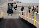 A forklift sits at the base of a ramp leading upwards. a person is sitting inside the cab of the forklift. Seven people stand off to one side and are looking at the forklift.