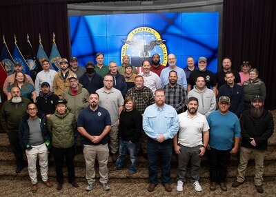 Photo of 34 men and women in working cloths standing on steps inside of a conference room with DLA Disposition Services' command logo on a screen behind them