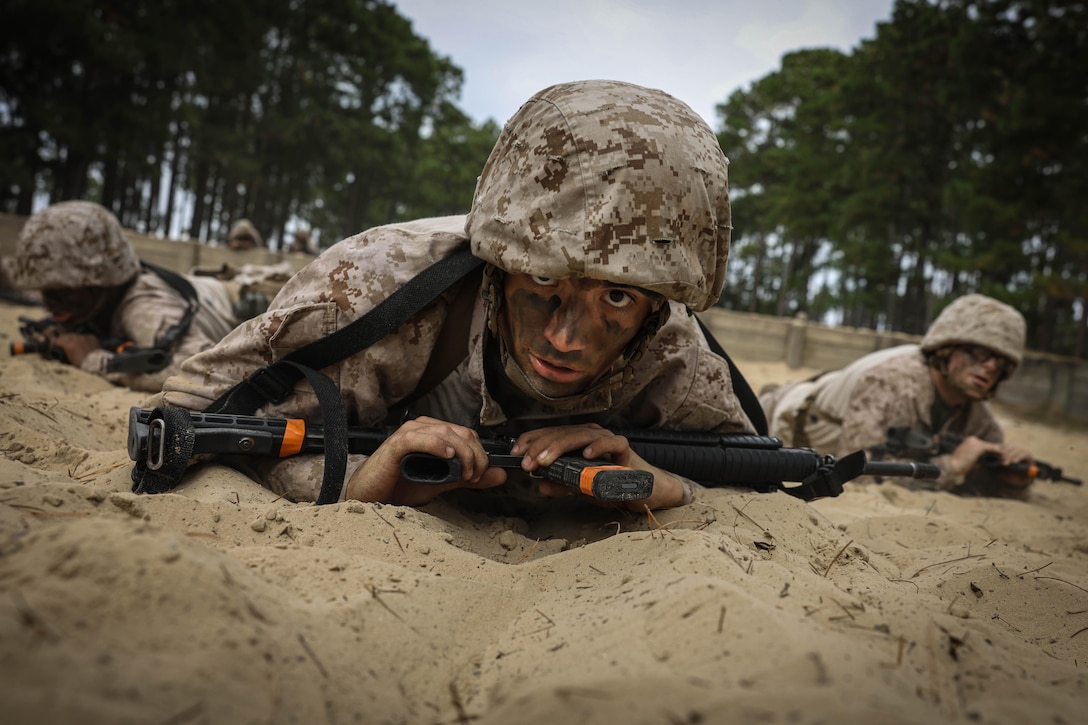 Uniformed recruits crawls through sand while holding weapons.