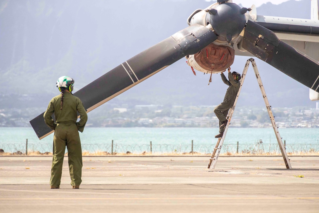 A Marine observes another Marine standing on a ladder while working on an aircraft propeller.