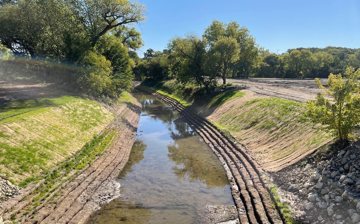 A creek runs between two creek banks with grass on either side, trees and a blue sky in the background.