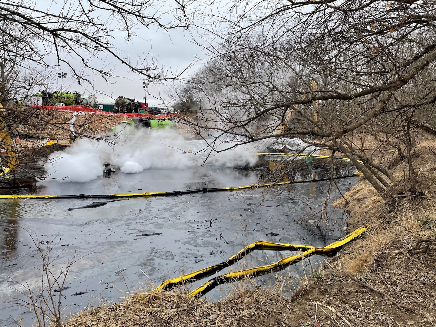 Black oil is seen in a creek with trees on the right and construction equipment in the background.