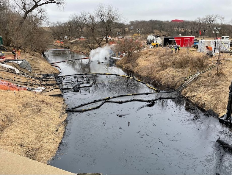Black oil is seen in a stream between two stream banks with construction equipment to the right.