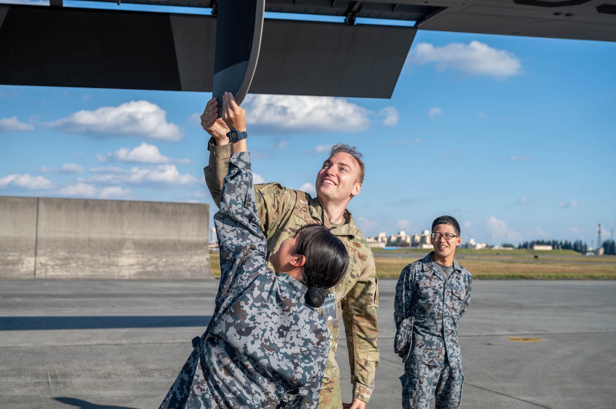 Three people in uniform are in the photo. Two of them are interacting with the propeller of an aircraft while the other person stand adjacent to them watching and smiling.