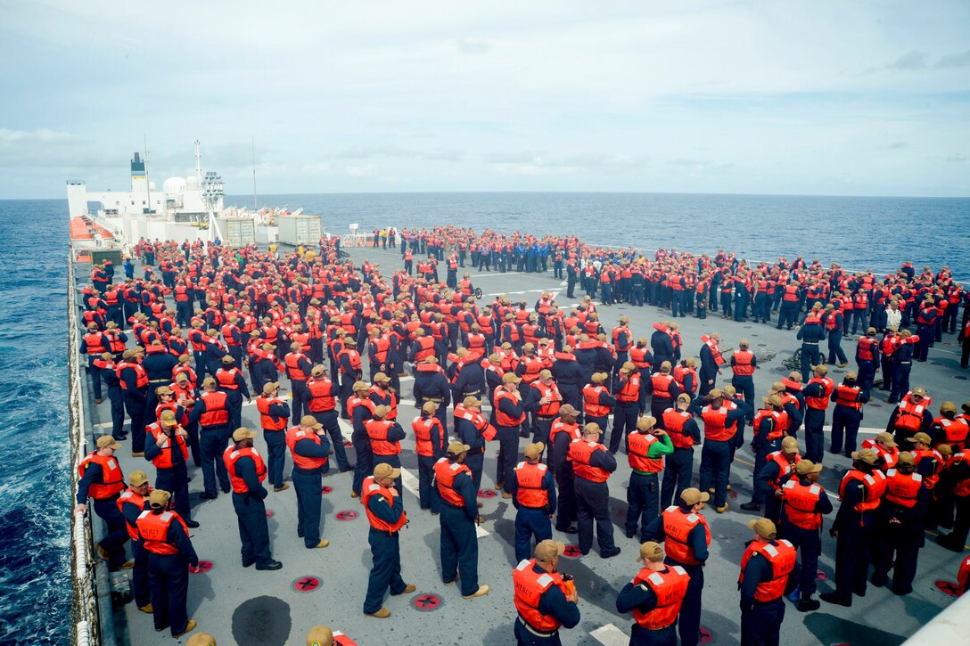 Dozens of staff members wearing orange life vests gather on the deck of a ship while transiting a body of water.